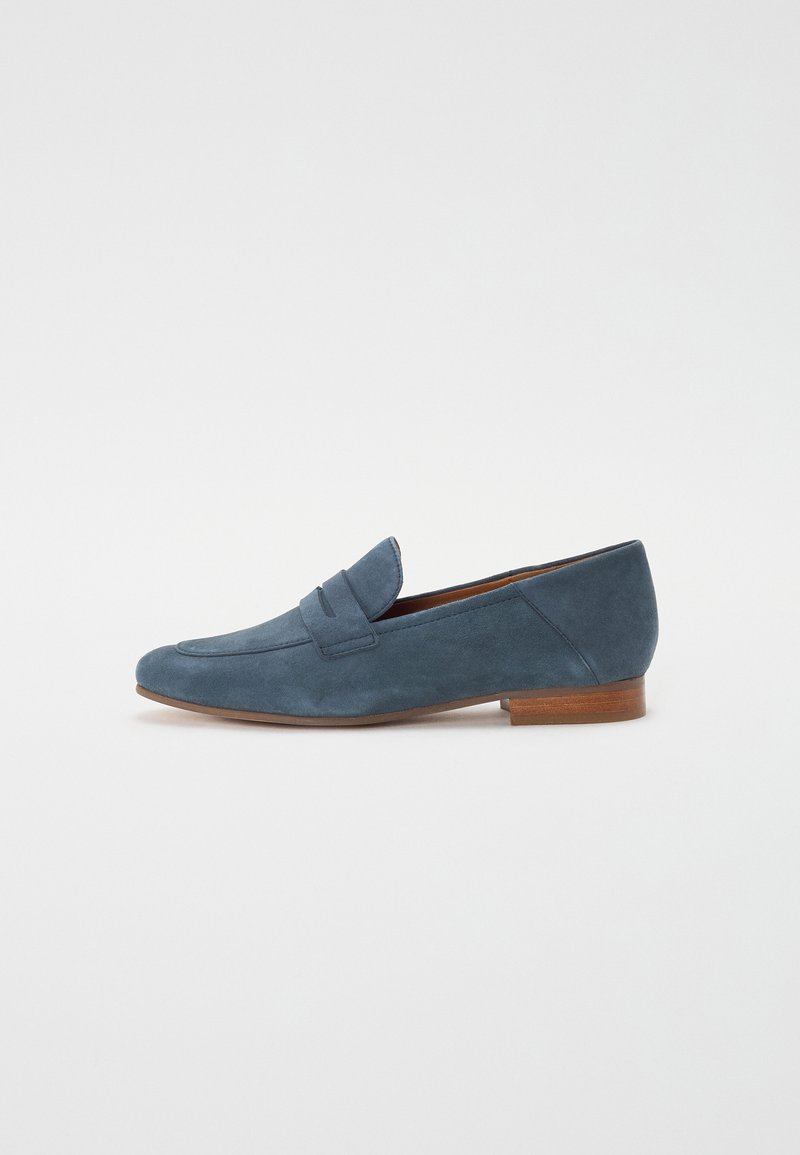 Mocassins en suede bleu avec un talon plat, présentant un design cousu de style mocassin et un détail de bandeau avant. Texture lisse avec une forme minimaliste.