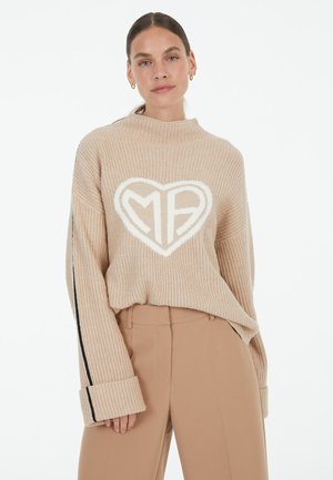 Beige knitted sweater with high neckline and oversized sleeves, featuring a white heart-shaped design with "MB" inside. Black side stripes.