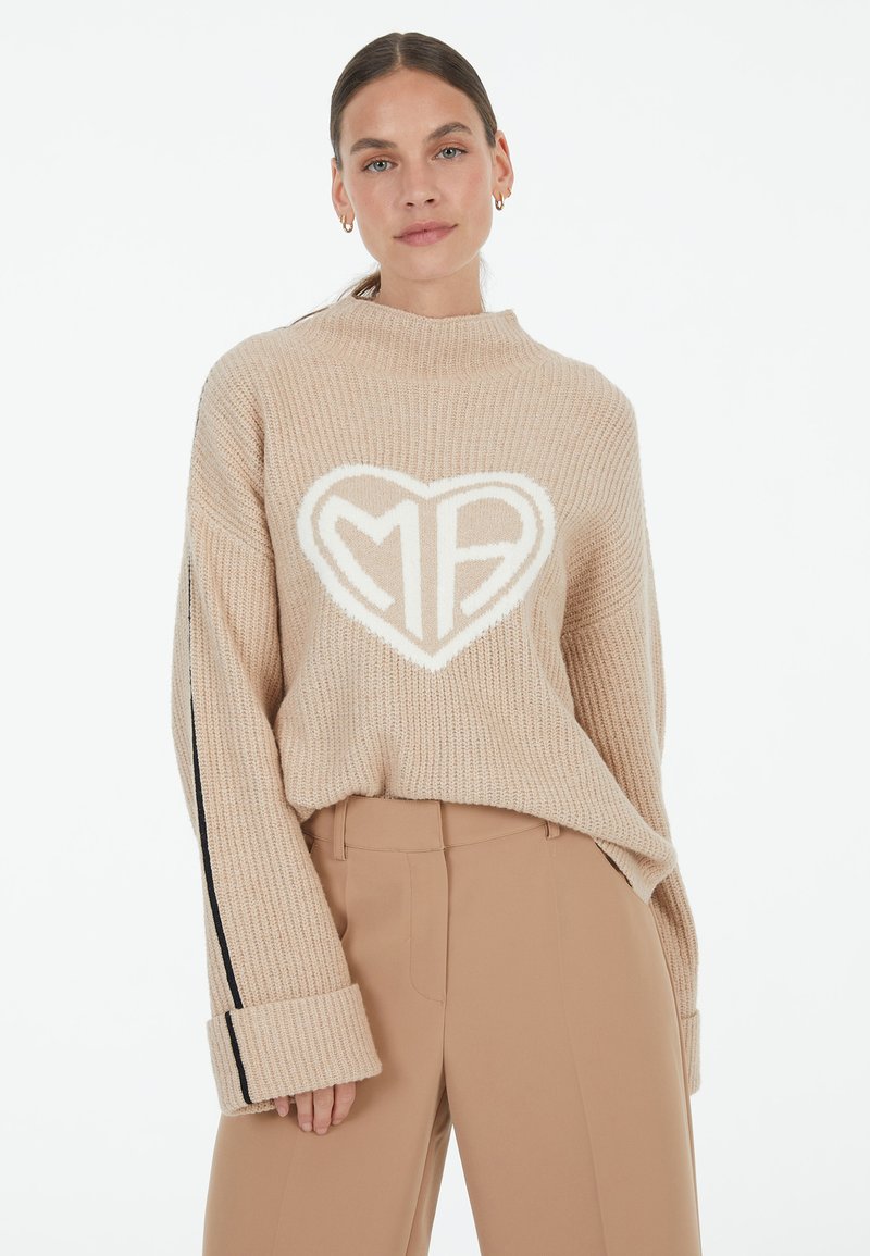 Beige knitted sweater with high neckline and oversized sleeves, featuring a white heart-shaped design with "MB" inside. Black side stripes.