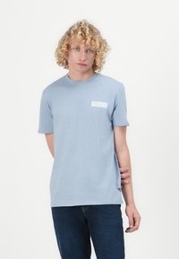 Helles blaues T-Shirt mit kurzen Ärmeln, das ein kleines weißes Logo auf der linken Brust, einen Rundhalsausschnitt und eine weiche Stoffstruktur aufweist.
