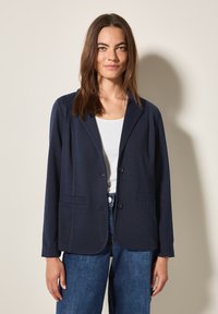 Blazer bleu marine en tissu doux, avec un col cranté, deux poches avant et fermeture à boutons. Associé à un haut blanc.