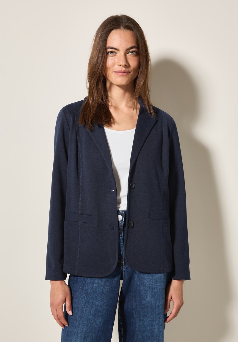 Blazer bleu marine en tissu doux, avec un col cranté, deux poches avant et fermeture à boutons. Associé à un haut blanc.