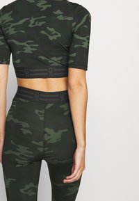 Lyhythihainen, tummanvihreä camo-kuosiset crop-top ja legginsit. Varustettu mustalla elastisella vyötärönauha, jossa on logopainatus.