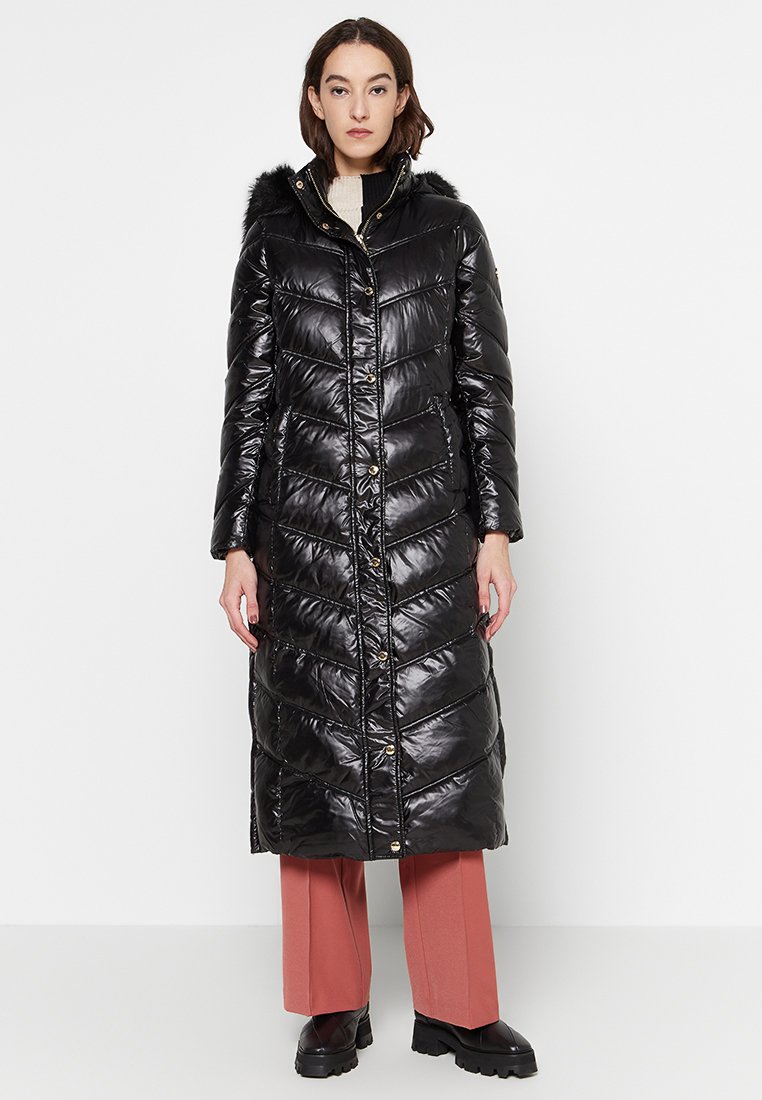 Donna che indossa un lungo cappotto nero lucido imbottito con cappuccio e pantaloni rosa a gamba larga, in piedi su uno sfondo bianco.