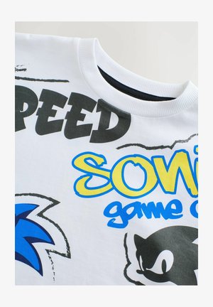 T-shirt bianco in cotone con testo blu e giallo che recita "Sonic", dettagli neri e grafiche stilizzate dei personaggi. Collo a girocollo rigato.