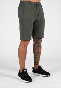 Groene katoenen sportshorts met zijzakken, elastische tailleband en trekkoord. Zwarte sportschoenen met witte zolen en een gestructureerd oppervlak.