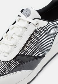 Baskets blanches et noires Michael Kors avec lacets blancs et détail de clous argentés sur le côté, nom de la marque visible.