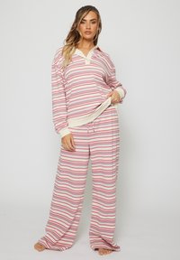 Kvinna som står barfota iförd en långärmad topp med rosa, vit och grå ränder samt matchande vida byxor mot en enfärgad bakgrund.