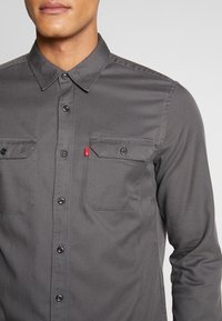 Grå långärmad skjorta med knäppta fickor, spetsig krage och en liten röd logotyp på vänster sida. Enfärgad strukturerad tyg.