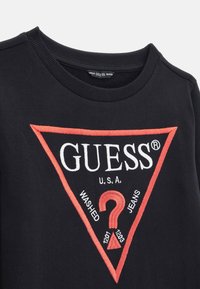 Sudadera de algodón negra con cuello redondo, que presenta un logo triangular rojo con la palabra "GUESS" y el texto bordado "WASHED JEANS" junto con números.