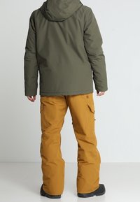 Zelená izolovaná bunda s kapucí, spárovaná s oranžovými cargo kalhotami s kapsami. Černé boty dokončují outfit připravený na venkovní aktivity.