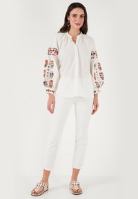 Witte blouse met kleurrijke bloemenborduursels op de opgeblazen mouwen, knoopsluiting aan de voorkant, gecombineerd met witte slim-fit broek en sandalen.