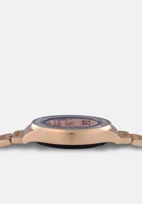 Orologio dorato con cassa tondeggiante, cinturino testurizzato e quadrante trasparente con numeri arabi e indicatori a punto. Corona distintiva sul lato.