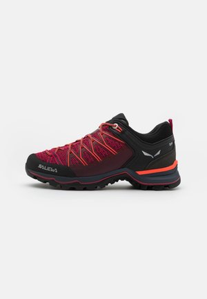 Chaussure de randonnée basse rouge et noire avec une semelle robuste, des lacets rouges et des logos de marque sur le côté et le talon, isolée sur un fond blanc.