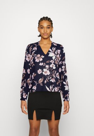 Blusa florido azul-marinho com mangas longas e decote em V, apresentando padrões florais em rosa-claro e branco. Usada com uma saia mini preta.