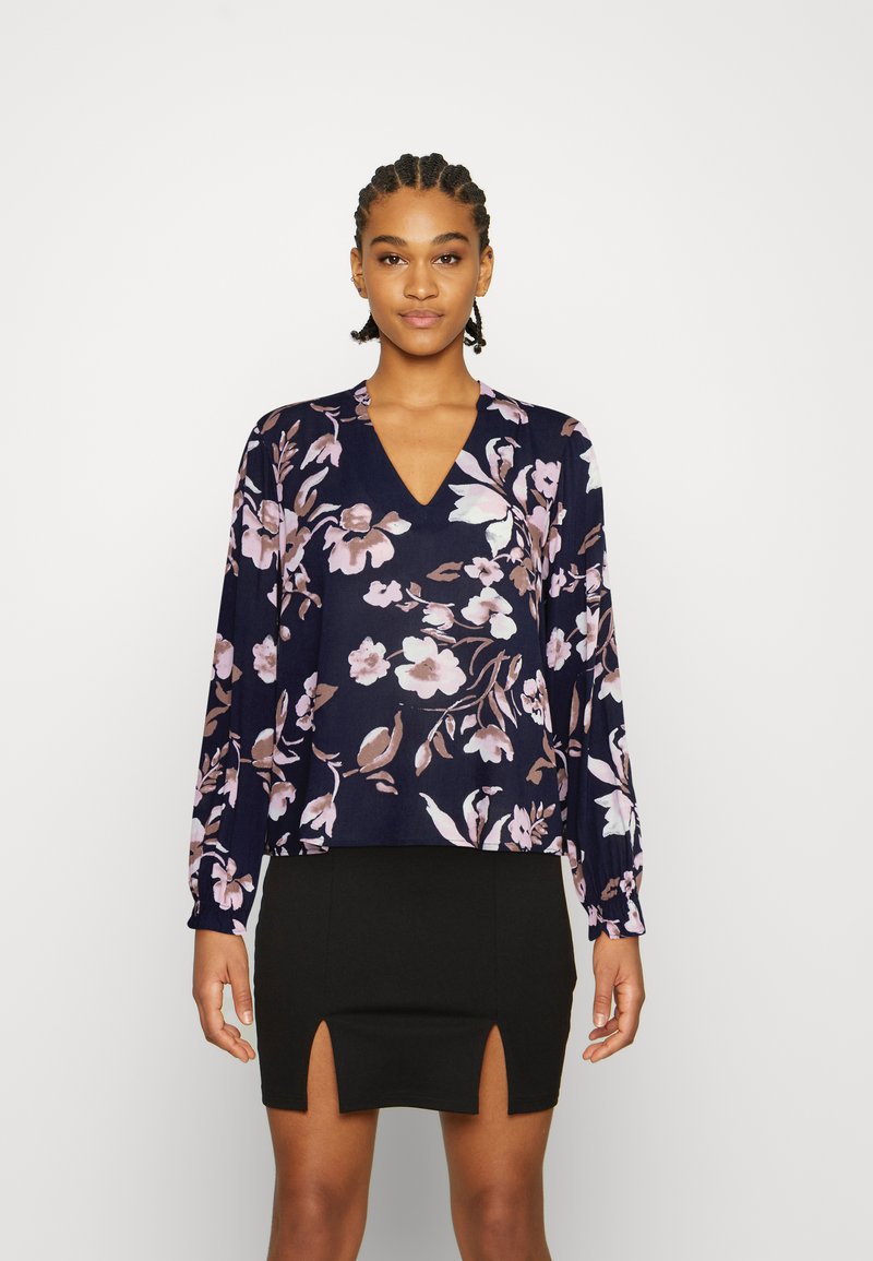 Blusa florido azul-marinho com mangas longas e decote em V, apresentando padrões florais em rosa-claro e branco. Usada com uma saia mini preta.