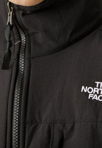 Zwarte geïsoleerde jas met een hoge kraag, ritssluiting aan de voorkant en geborduurd logo. Het materiaal lijkt glad met een matte afwerking.