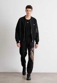 Black bomber jacket with zip closure, paired with black sweatpants featuring a painted design in earthy tones. Black sneakers complete the outfit.