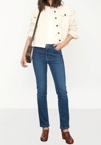 Světlá béžová manšestrová bunda s zapínáním na knoflíky, doplněná tmavě modrými úzkými džínami. Hnědé dřeváky a malá hnědá kabelka na rameno dokončují vzhled.