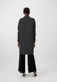 Manteau long gris foncé avec un col, à coupe droite et manches longues. Le modèle porte un pantalon noir à jambes larges et des chaussures à talons.