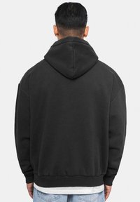 Musta kapuutsiga sweatshirt, millel on lõdva istuvusega lõige, varustatud kangaroo tasku ja soonikuga varrukatega, valmistatud pehmest kangast. Kapuutsi peal on nähtavad õmblused.