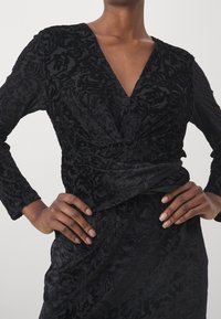 Robe noire avec une texture florale, décolleté en V profond et manches longues, présentant un design avant torsadé et une silhouette ajustée.