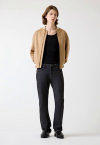Veste bomber beige à texture lisse, portée sur un débardeur noir. Associée à un pantalon en jean foncé et des chaussures noires. Design simple et moderne.