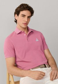 Jeune homme assis, portant un polo rose avec un petit logo et un pantalon blanc, ceinture tressée beige, fond gris neutre.