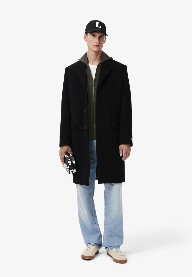 Zwarte wollen jas met een geknipt kraag, gecombineerd met een olijfgroene hoodie en lichtblauwe spijkerbroek. Model houdt een gestructureerde tas vast.