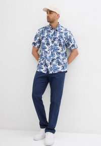 Camicia a maniche corte, blu e bianca con motivo floreale, abbinata a pantaloni navy e sneakers bianche. La camicia presenta un motivo di foglie tropicali.