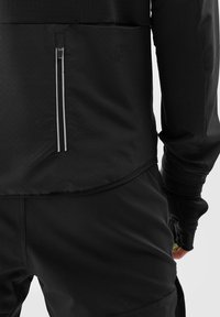 Zwarte sportkleding met een ritszak, gestructureerde stof, nauwsluitende pasvorm en lange mouwen met duimgaten voor extra bedekking.