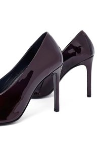 Une paire de chaussures à talons hauts vernies de couleur bordeaux foncé avec des bouts pointus et des talons aiguilles sur un fond blanc.