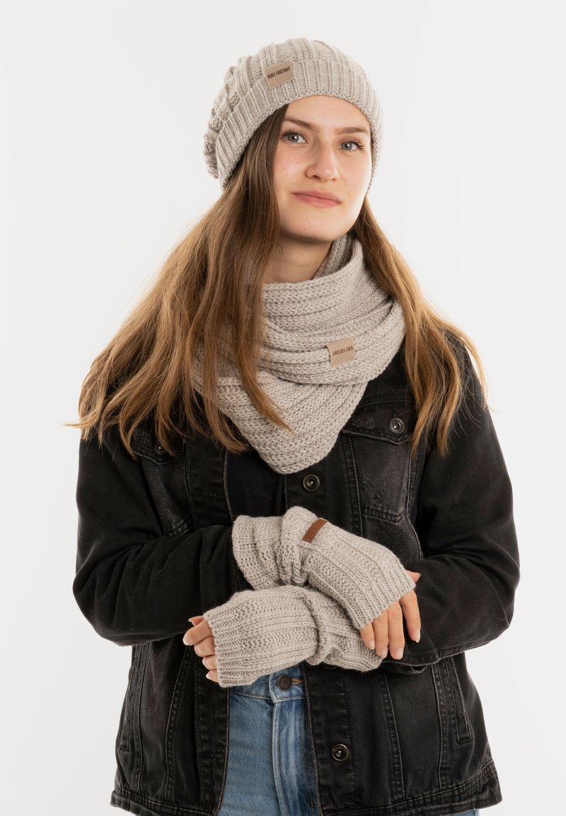 Gebreide beanie, sjaal en vingerloze handschoenen in lichtgrijs, met een geribbelde textuur en leren logoaccenten tegen een donkere jas.