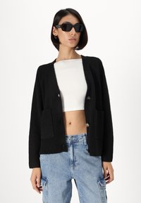Cardigan nero con polsini a costine, due tasche frontali e bottoni tondi. Indossato sopra una maglietta crop bianca e abbinato a jeans azzurri chiari.