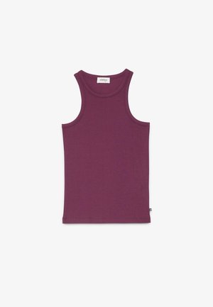 Vrouw staat in een eenvoudige kamer met een mouwloze paarse top, gestreepte uitlopende broek, een zwarte riem met studs en zilveren spitse schoenen.