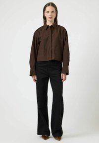 Camicia marrone ritagliata con colletto a punta, due tasche frontali e maniche lunghe, abbinata a pantaloni neri svasati e scarpe marroni a punta.