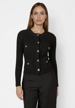 Frau trägt eine eng anliegende schwarze, gerippte Strickjacke mit silbernen Knöpfen und vier Klappentaschen, kombiniert mit schwarzer Hose vor einem schlichten Hintergrund.