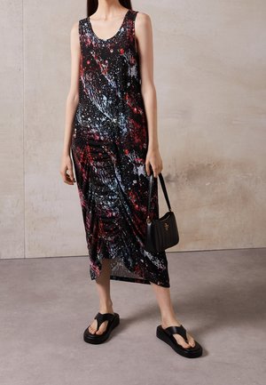 Vrouw draagt een mouwloze zwarte jurk met rode, blauwe en witte spetterpatronen, houdt een zwarte handtas vast en draagt zwarte plateauzandalen.