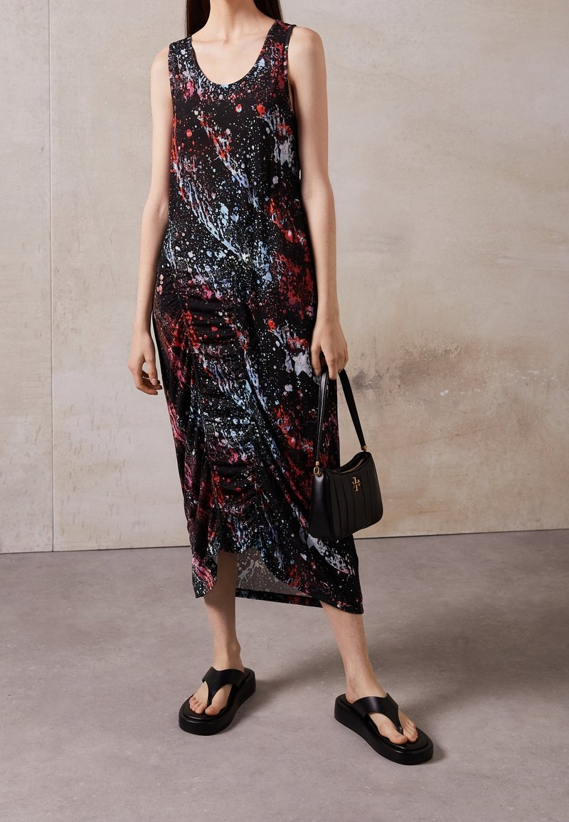 Kvinde iført en ærmeløs sort kjole med røde, blå og hvide sprøjtemønstre, der holder en sort håndtaske og bærer sorte plateau-sandaler.