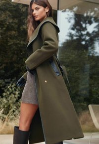 Alyvuogių žalios spalvos paltas su dideliais atvartais, juodais odiniais akcentais ir surišta diržu. Derinamas su pilku sijonu ir juodomis pirštinėmis, pristatytas lauke.