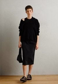 Zwarte oversized gebreide trui met een open schouder, gecombineerd met een strakke zwarte midi-rok, sandalen met dikke banden en een zwarte handtas.