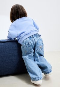 Világoskék pulóver bordázott mandzsettákkal, laza világos farmernadrággal, amelyen hímzett foltok vannak a hátsó zsebeknél. Fehér tornacipő.