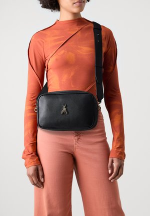 Schwarze Leder-Crossbody-Tasche in rechteckiger Form, mit metallischem Insektenlogo und verstellbarem Riemen; getragen mit einem orangefarbenen Langarmoberteil und pinken Hosen.