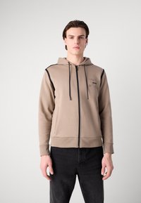 Haut à capuche beige zippé avec un cordon de serrage, accents noirs sur les épaules et poches latérales. Le tissu semble lisse et extensible.