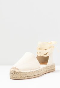 Chaussure espadrille en toile beige clair avec une semelle en jute, dotée d'un bout rond et d'un long ruban jaune pour nouer autour de la cheville.
