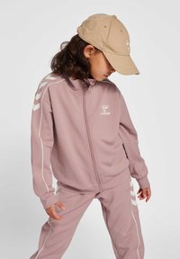 Ljusrosa träningsdräkt med vita detaljer och logotyp. Jacka med blixtlås och byxor med elastisk midja. Beigefärgad keps fulländar looken.