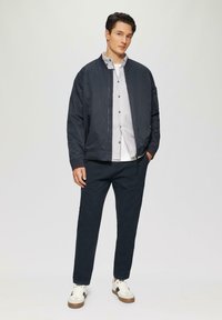 Marine bomberjack over een wit en marine gestreept shirt, gecombineerd met marinebroeken en witte sneakers met bruine accenten. Eenvoudig ontwerp, gladde stof.