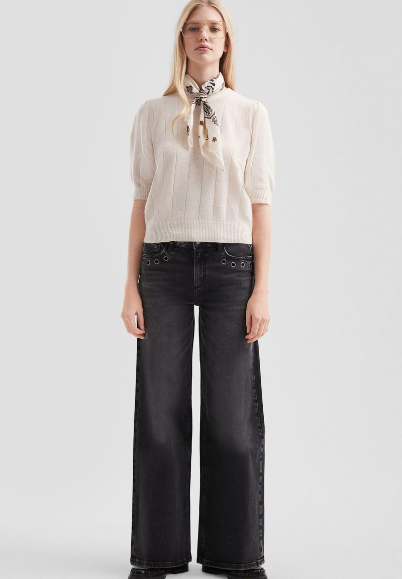 Crème gekamde gebreide top met korte mouwen, gecombineerd met hoog-waist zwarte wijde jeans. Sjaal geknoopt om de nek met florale print en hardware accenten.