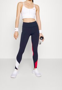 Vit sport-bh med Nike-logotyp, högmidjade marinblå leggings med röd och vit färgblockaccent, ihop med vita träningsskor.