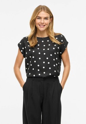 Smilende kvinne med blondt hår har på seg en svart kortermet bluse med hvite prikker og svarte høytsittende bukser, hender i lommer.